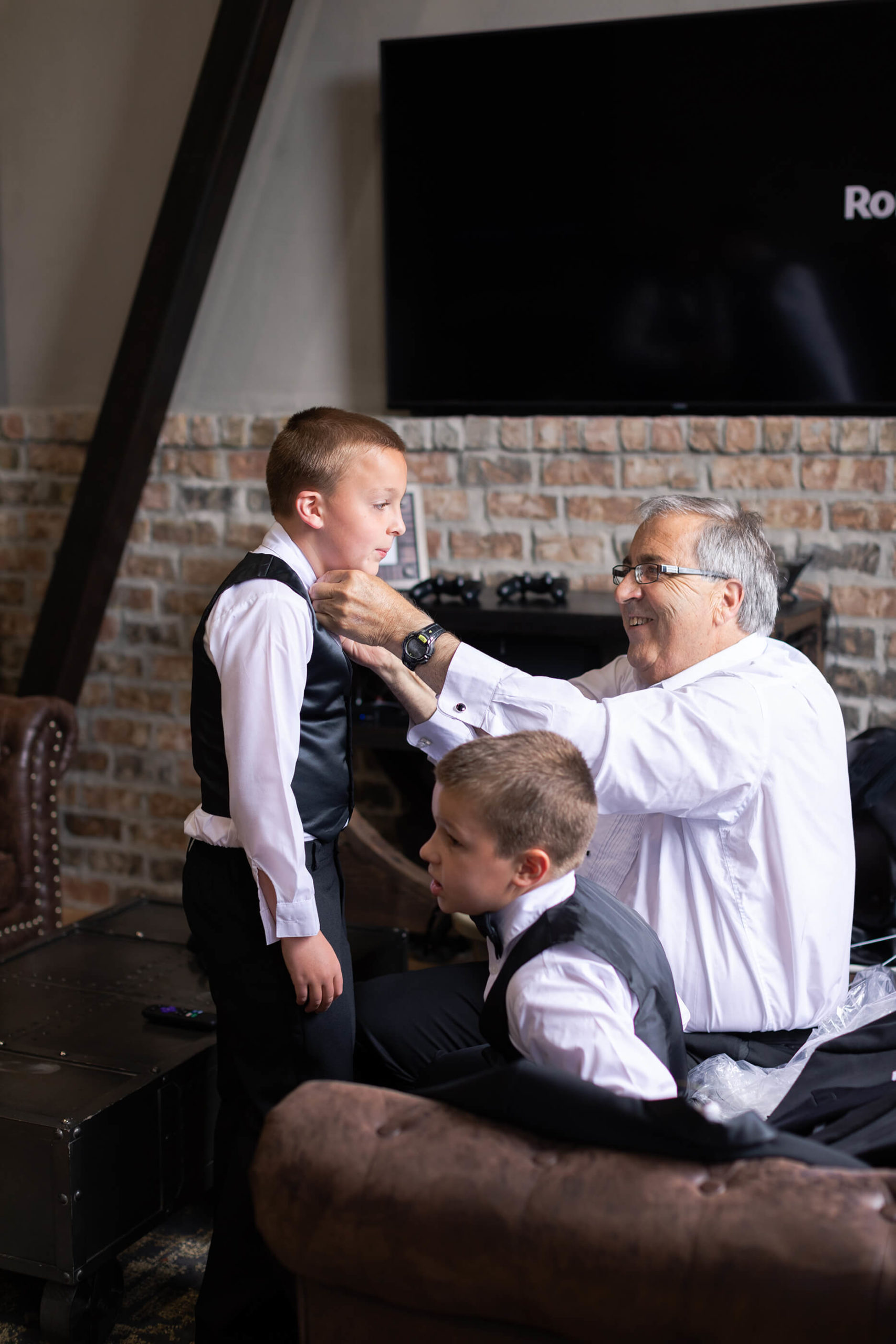 grandfather ties tie on grandson on wedding day bride looks at herself as she gets ready on her wedding day reception space at Iron Manor in Houston Texas by Swish and Click Photography