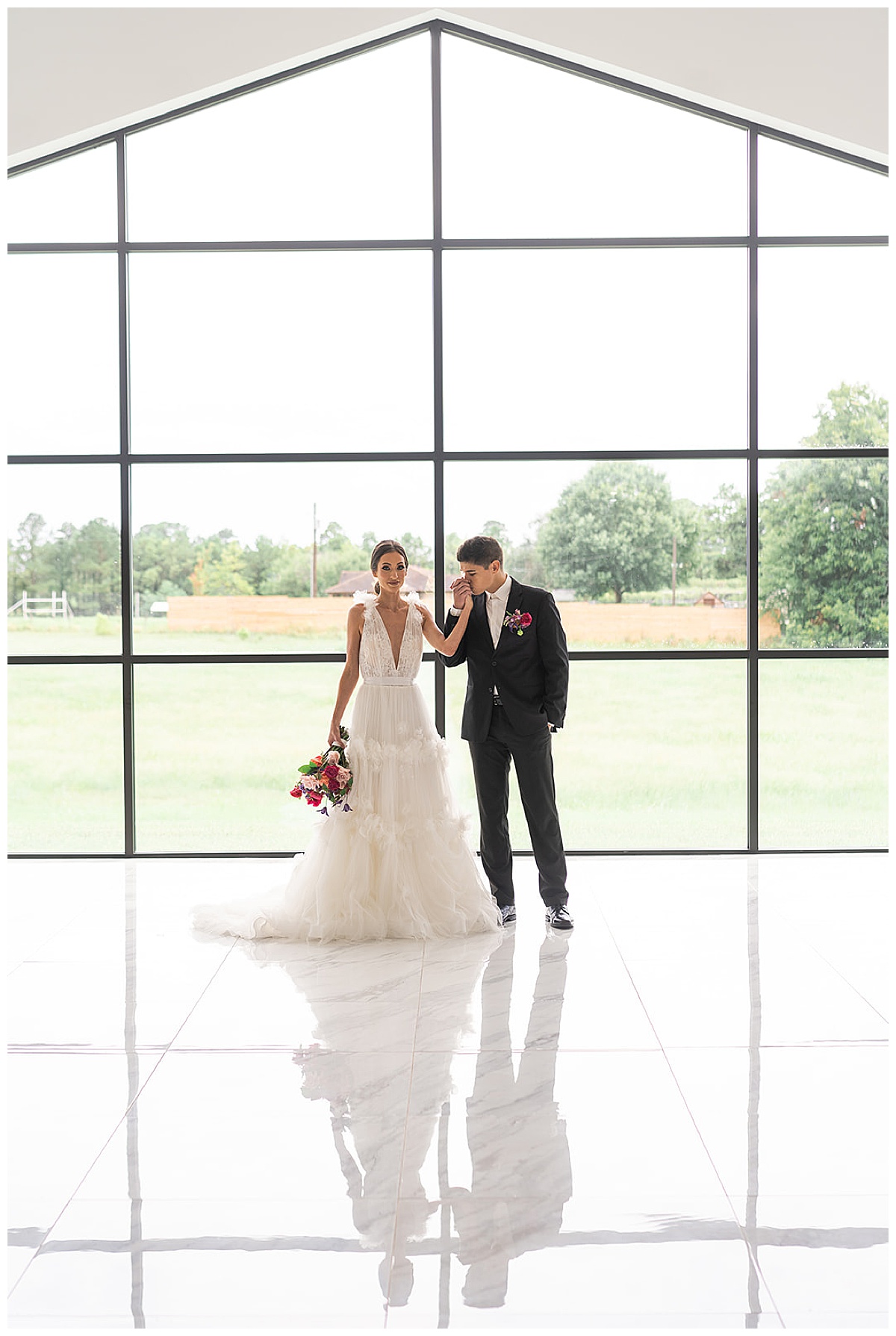 Groom kisses bride's hand in front of large windows at The Homestead