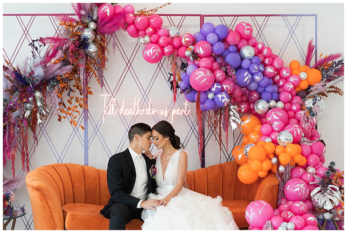 Couple sit together underneath colorful balloon arch showing off 70's Inspired Wedding Ideas
