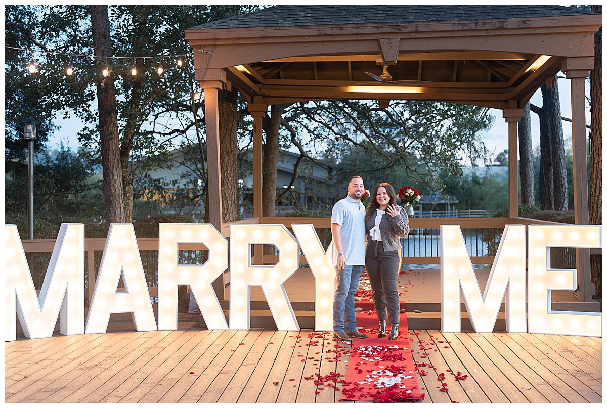 Engaged couple standing in front of letters spelling Marry Me