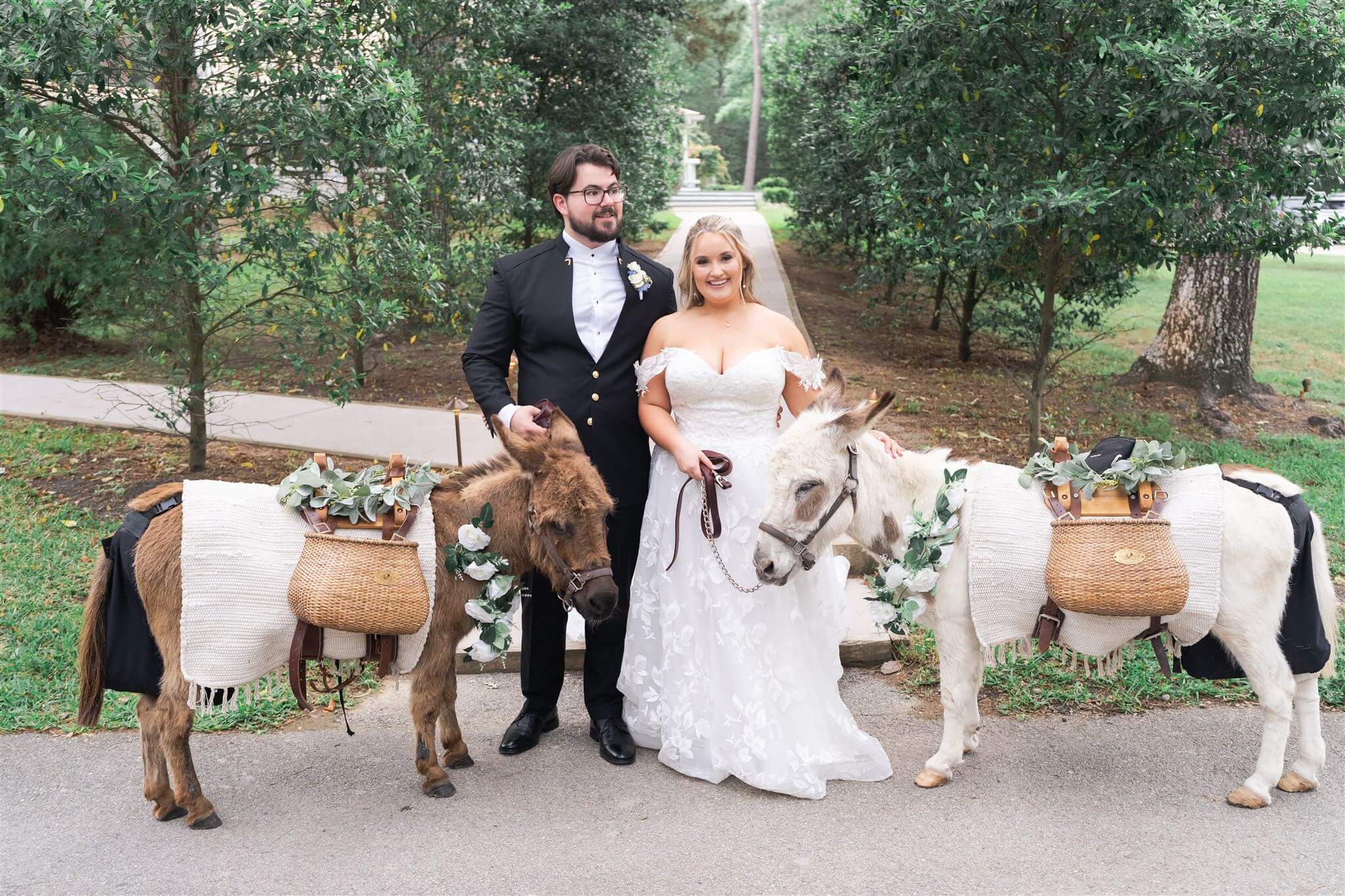 A curvy bride faces the camera with her ankles crossed to get an S shape with her groom standing next two her and two donkeys.