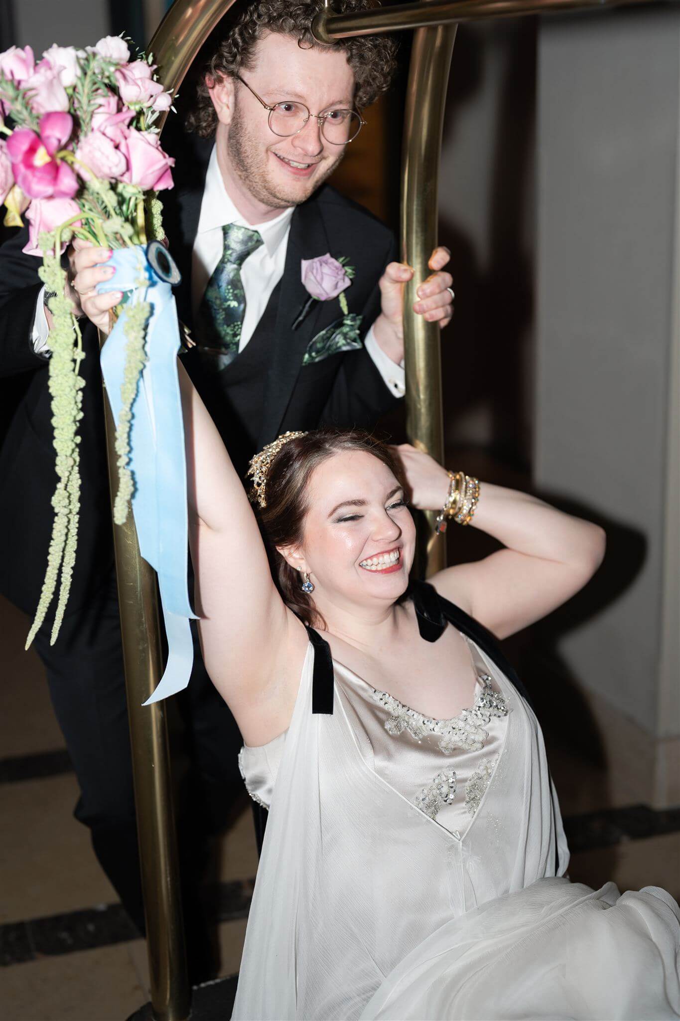 A curvy bride with her bouquet in the air being pushed on a hotel luggage cart by her groom!